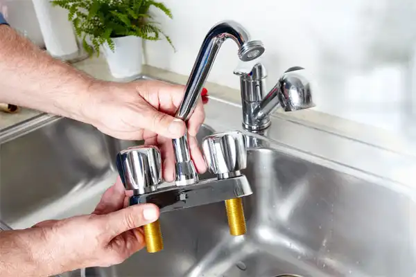 Hands holding a detached kitchen faucet with two knobs, above a stainless steel sink.