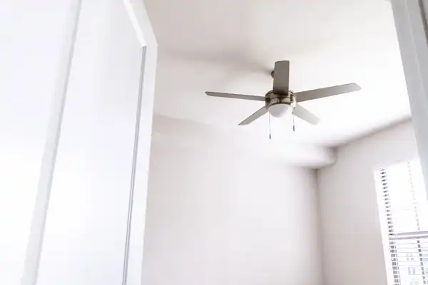 A white ceiling fan with four blades is mounted on a white ceiling in a room with white walls and a partially visible window with blinds.