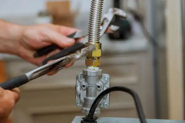 Hands using pliers to tighten a brass fitting on a flexible gas line connected to a metal valve.