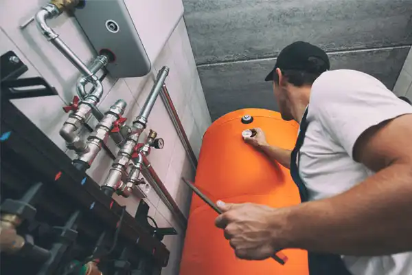 A technician in a cap adjusts controls on a large orange water heater in a utility room, surrounded by pipes and valves.