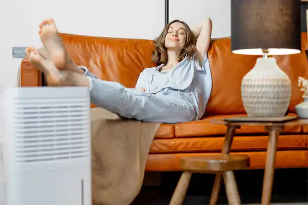 A woman in casual clothes relaxes on an orange sofa with her feet up, near a black lamp and a dehumidifier.
