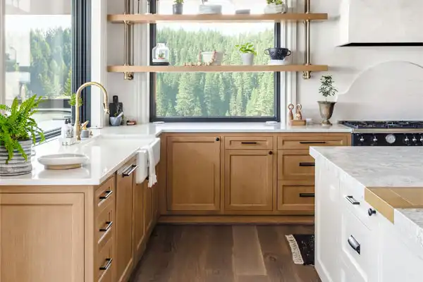 Modern kitchen with wooden cabinets, white countertops, and open shelves. Large window with a view of greenery. Contains potted plants, sink, and stove.