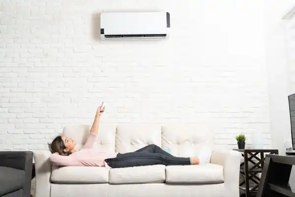 A person relaxes on a sofa, pointing a remote control at a wall-mounted air conditioner.