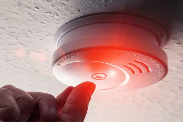 A person pressing a button on a ceiling-mounted smoke detector emitting a red light.