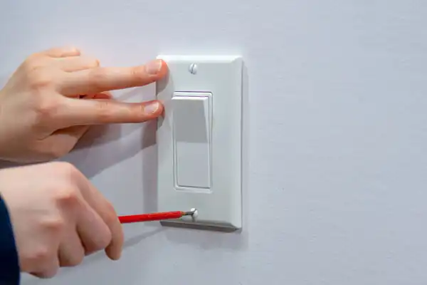 Person using a screwdriver to install or remove a white light switch cover on a wall.