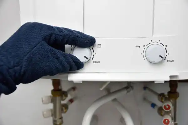 Gloved hand adjusting the temperature dial on a water heater, with visible pipes and fittings below.