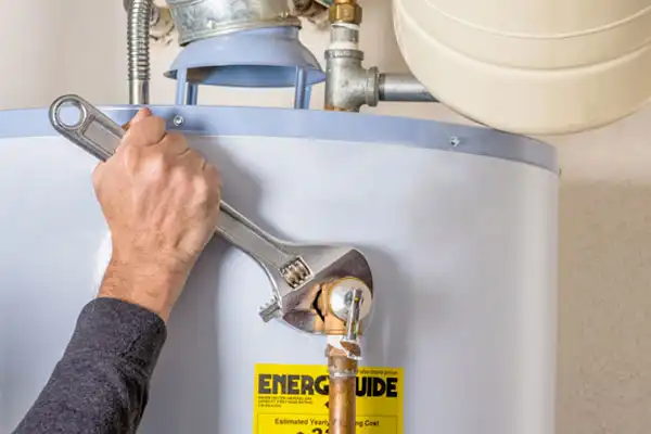 A person uses a wrench to tighten a pipe on a water heater, with an EnergyGuide label visible on the appliance.