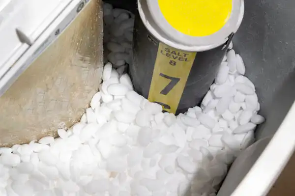 Salt pellets are scattered around a yellow tube marked with salt levels inside a water softener tank.