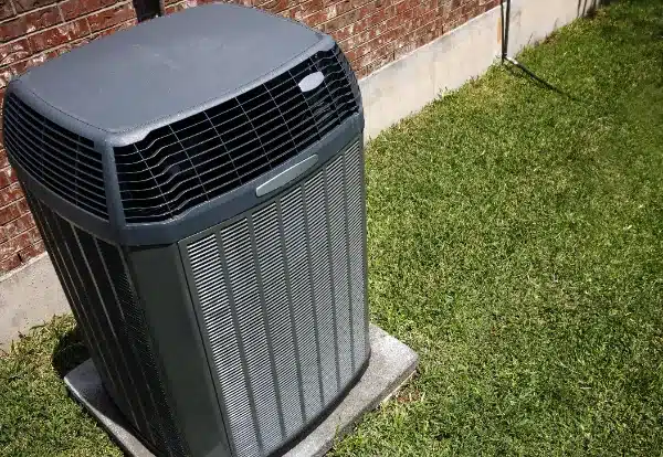 Outdoor air conditioning unit on a concrete slab sits next to a brick wall surrounded by grass.
