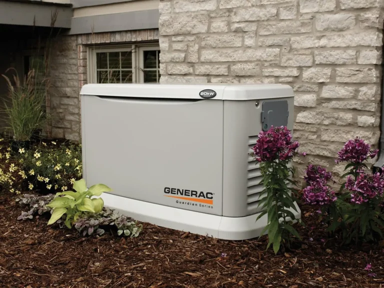 A Generac Guardian Series backup generator is installed outside a brick house, surrounded by purple flowers and small shrubs.