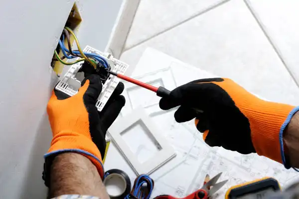 Person wearing orange gloves using a screwdriver to install or repair an electrical outlet on a wall, with tools and materials nearby.