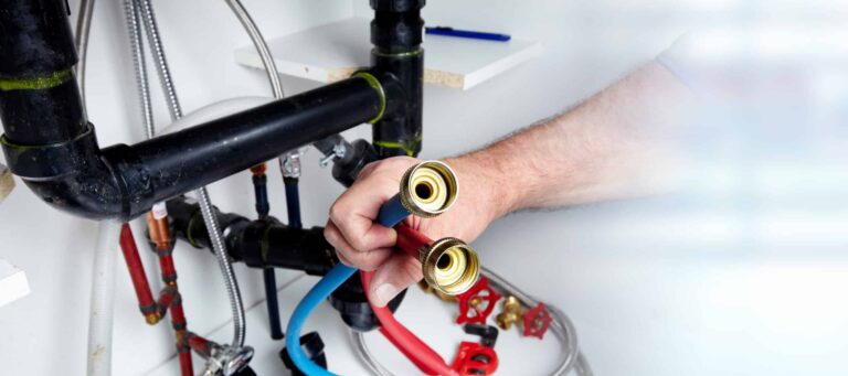 A person connects colored hoses to plumbing pipes under a sink or cabinet.