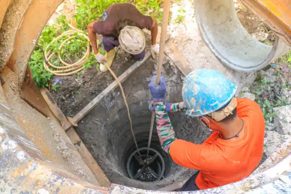 Two workers wearing helmets lower equipment into a deep, circular well using a rope. One is in orange and blue, the other in a dark shirt.